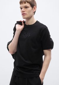 Fekete pamut póló tónusos "BOSS" logóval. A modell egy fekete táskát tart, az egyszerű kerek nyakkivágású és laza szabású pólót visel.