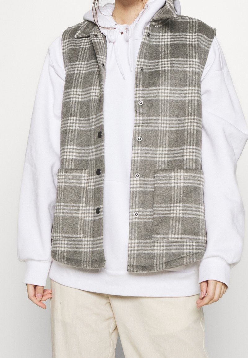 Personne portant un gilet boutonné à carreaux gris et blancs sur un sweat à capuche blanc et un pantalon crème, les mains détendues sur les côtés.