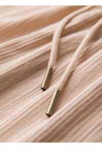 Gros plan sur un tissu beige à texture côtelée verticale et deux cordons beiges avec embouts métalliques.