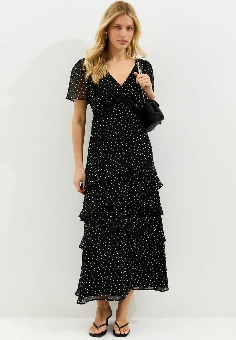 Femme blonde portant une robe midi noire à pois blancs, manches courtes en tulle, jupe à volants, sac noir et sandales à talons noires.