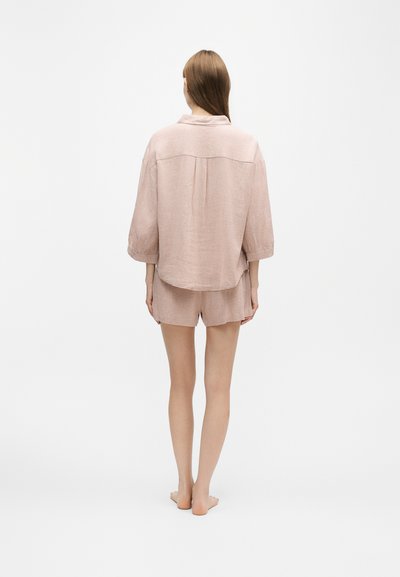 Γυναίκα στεκόμενη ξυπόλητη με την πλάτη, φορεί χαλαρό απαλό ροζ υφασμάτινο σακάκι με μανίκια τριών τετάρτων και ταιριαστά σορτς.