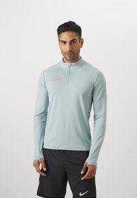 Langærmet, lys blå atletisk top med lynlås i kraven, med et pink Nike-logo på brystet og tommelhuller i ærmerne.