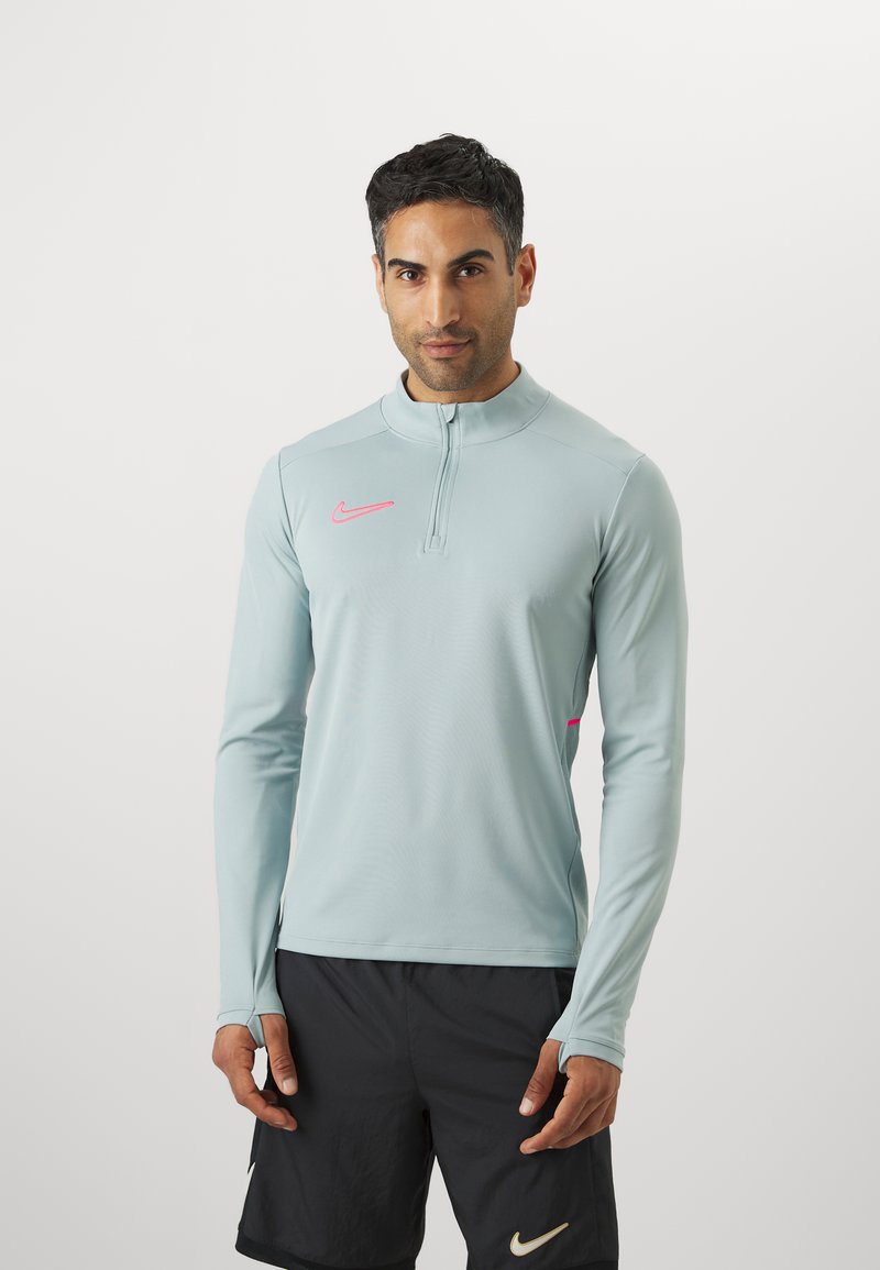 Langærmet, lys blå atletisk top med lynlås i kraven, med et pink Nike-logo på brystet og tommelhuller i ærmerne.