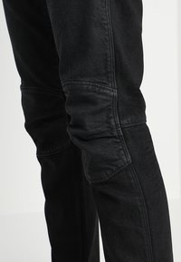 Mustat farkut, joissa on kapeneva malli, joissa on erottuvat polviosat, tekstuuripinta ja hienovaraiset ompeluotteet sivusaumoissa.