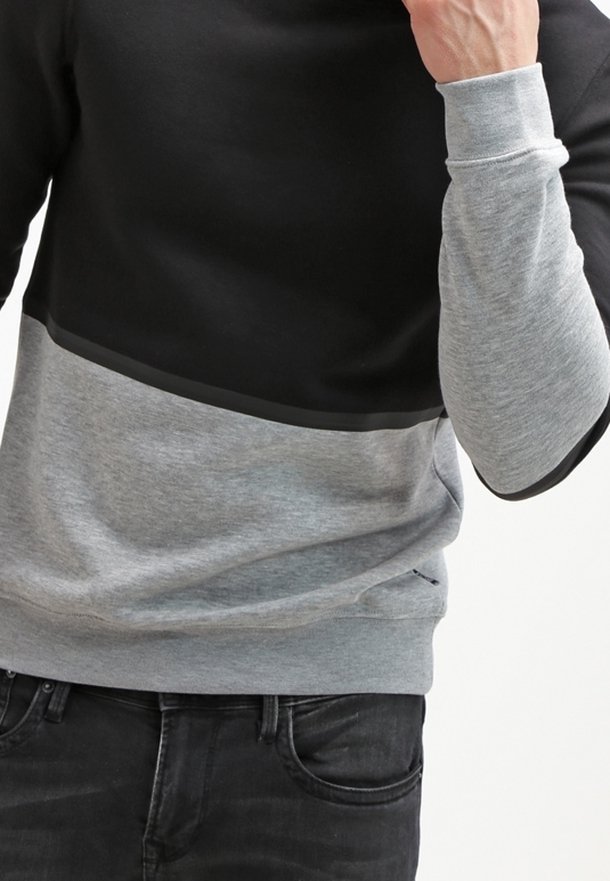 Hombre que lleva puesta una sudadera bloqueada en negro y gris con mangas ajustadas, combinada con jeans de mezclilla negros.