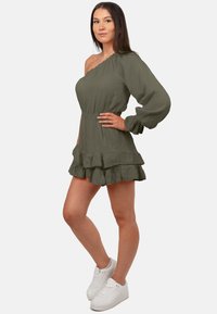 Robe olive verte à une épaule en tissu texturé avec une taille cintrée et un ourlet volanté, associée à des baskets blanches.