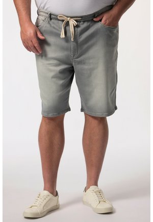 Uomo che indossa pantaloncini grigi con coulisse e scarpe da ginnastica bianche, in piedi con le mani vicino alle tasche su uno sfondo uniforme.