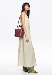 Bordowa skórzana torebka z podwójnymi uchwytami i regulowanym paskiem. Modelka ubrana w kremową sukienkę z falbanami i oliwkowo zielone spodnie.