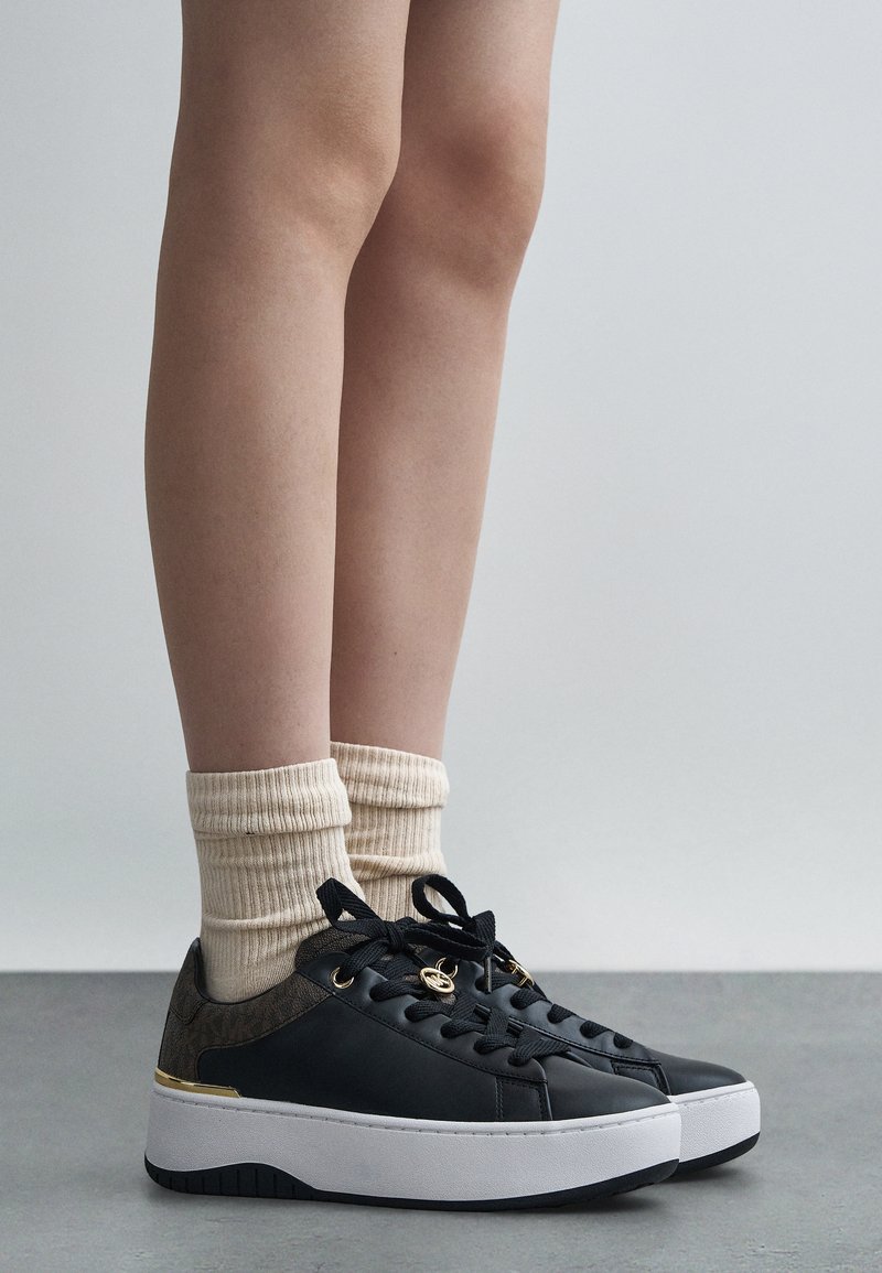 Baskets compensées noires avec un accent marron texturé, semelle en caoutchouc blanche et lacets noirs, assorties à des chaussettes crème côtelées.