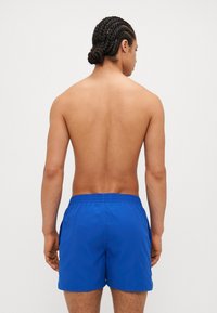 Blå svømmebukser med elastisk talje og sidelommer, glat tekstur. Modellen vises fra ryggen.