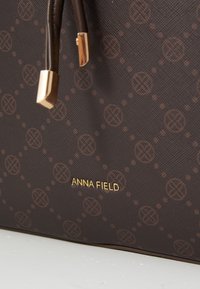 Sac à main marron à motifs avec une surface texturée, des accents en métal doré et des poignées en cuir avec des extrémités métalliques ; logo "ANNA FIELD".