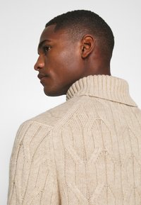 Suéter de punto cable beige con un alto cuello acanalado, que presenta un patrón texturizado y un tejido grueso. El diseño es ajustado en el cuello.