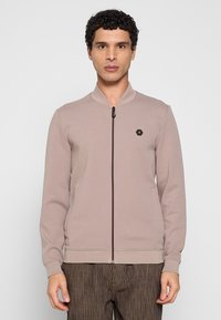 Lichtbruine sweatvest met rits en geribde kraag, met een klein zwart zeshoekig logo op de borst en een gladde stoftextuur.