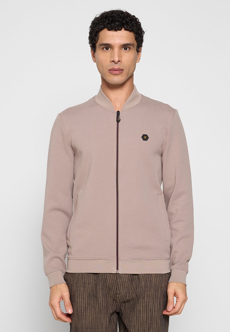 Lichtbruine sweatvest met rits en geribde kraag, met een klein zwart zeshoekig logo op de borst en een gladde stoftextuur.