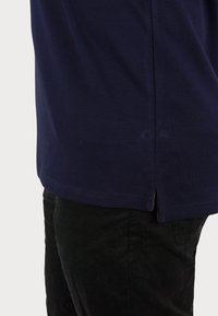 Polo shirt blu navy con tessuto a trama, presenta una fessura laterale all'orlo ed è indossato con pantaloni di velluto a coste neri.