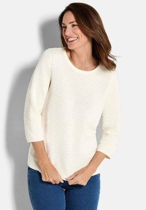 Lächelnde Frau mit braunen Haaren, die einen strukturierten weißen Pullover mit drei Viertel-Ärmeln und blaue Jeans trägt, vor einem hellen Hintergrund.