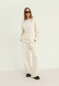 Mulher vestindo um sweatshirt oversized em creme e calças largas, óculos de sol pretos e sapatos pretos, de pé com as mãos nos bolsos.