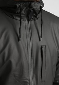 Närbild av en svart vattentät jacka med centralsida dragkedja, snörning i huvan och lodrät dragkedjeficka på bröstet.