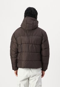 Brun pufferjakke med hætte, der har vandrette quiltede sektioner og en glat tekstur. Bemærkelsesværdigt design inkluderer en rundet hætte og kort længde.