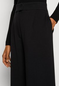 Pantalon noir à jambes larges avec une texture lisse, doté d'une taille ceinturée, de poches latérales et d'une coupe ajustée autour des hanches.