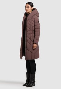 Barna párnázott hosszú kabát széles kapucnival, steppelt mintával, oldalzsebekkel és fekete cipzárral díszítve. Fekete leggings és csizma fölött viselve.