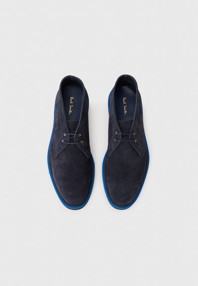 Chaussures en suede bleu marine avec des semelles en caoutchouc bleu et des œillets dorés. Elles présentent un design à coupe basse et une texture lisse avec un détail minimal.