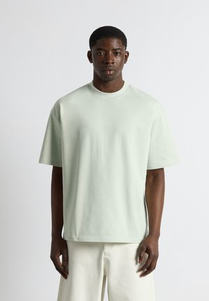 Jeune homme noir debout de face, portant un t-shirt oversize vert pastel clair et un pantalon crème sur un fond uni.