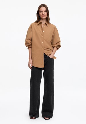 Femme debout avec une main dans la poche, portant une chemise ample couleur fauve boutonnée et un jean noir à jambes larges sur fond blanc.