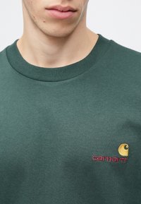 Zelená bavlněná mikina s krátkým rukávem a žebrovaným límcem, s vyšitým logem v žluté a růžové barvě na levé části hrudi.