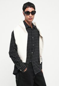 Czarna koszula w kratę z białym kołnierzykiem i rękawami, w połączeniu z kremowym swetrem w formie kamizelki. Czarna okulary w kształcie kocich oczu dopełniają stylizację.