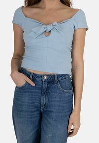 Donna che indossa un top a spalle scoperte a coste di un blu chiaro con un fiocco frontale e jeans a vita alta blu, con una mano in tasca.