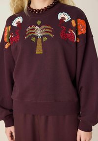 Sweatshirt met rode kat, groene palmboom en bloemenapplicaties van pailletten op bordeauxrode stof, gedragen met bijpassende bordeauxrode broek.