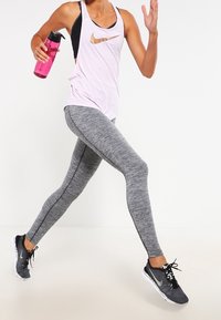 Světle růžový sportovní top s černými akcenty, šedé legíny s vzorem a černé sportovní boty. Drží růžovou láhev na vodu.