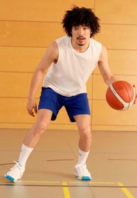 Vyrų atletas balta tank top ir mėlynomis šortais, driblinguodamas krepšinio kamuolį. Mūvi baltus sportbačius su spalvingais akcentais ant sporto salės grindų.