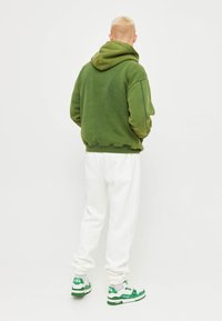 Persona con capelli biondi corti che indossa un hoodie verde, pantaloni bianchi e scarpe da ginnastica verde e bianche, in piedi con la schiena rivolta su uno sfondo semplice.