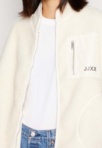 Kremowa kurtka polarowa z zamkiem błyskawicznym, z białą kieszenią na zamek i stylizowanym napisem "JJXX" z boku. Noszona na białej koszuli.