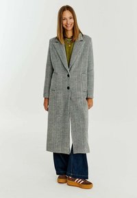 Šedobílý kabát se vzorem houndstooth s dvouřadým designem, velkými kapsami a klopami, spárovaný se zeleným topem a širokými džíny.