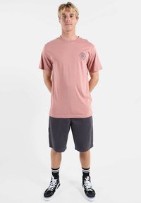 Jeune homme debout, les mains derrière le dos, portant un t-shirt rose poussiére, un short gris foncé, des chaussettes noires et des baskets montantes noires.