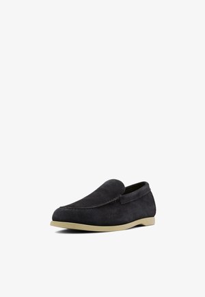 Mocassin en daim noir à enfiler avec semelle plate beige, bout arrondi et petit talon, présenté sur fond blanc.