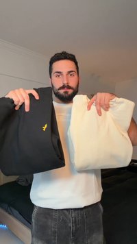 Mężczyzna z ciemnymi włosami i brodą trzyma w złożonych rękach czarną i białą bluzę, stojąc w pokoju z łóżkiem w tle.