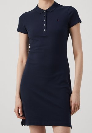 Femme portant une robe polo bleu marine ajustée à manches courtes avec patte de boutonnage et petit logo sur la poitrine, sur un fond uni.