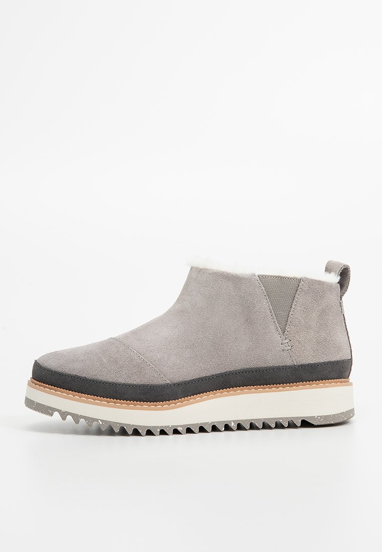Grå mocka ankelboot med vit pälsfoder, gummisula och texturerade grå accentuer. Har en rund tå och olika materialtexturer.