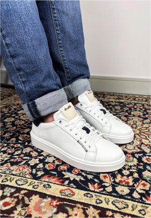 White leather sneakers with black accents worn with rolled-up blue jeans, standing on a multicolored floral patterned rug indoors.