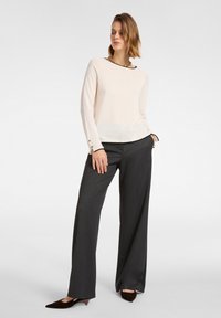 Helles beige Langarmoberteil mit schwarzem Besatz, seitlichen Knopfakzenten, kombiniert mit dunkelgrauen weiten Hosen und braunen Absatzschuhen.