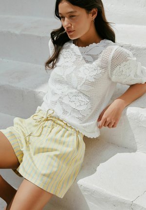 Naine istub valgetel treppidel, kandes valget lillelist pitsist pluusi ja kollaste triipudega paelaga lühikesi pükse.