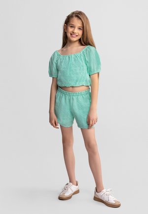 Młoda dziewczyna stoi i się uśmiecha, ubrana w miętowozieloną bluzkę z krótkim rękawem z ażurowego materiału oraz dopasowane szorty, na nogach ma białe trampki, na jednolitym tle.