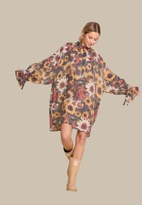 Blomstrete overdimensjonert kjole med lange ermer, med gule, oransje og hvite blomster på en mørk bakgrunn. Brukt med beigefargede gummistøvler.