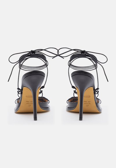 Μαύρα σανδάλια με ψηλά τακούνια και λεπτούς λουράκια στον αστράγαλο δεμένα με φιόγκους, από πίσω, σε λευκό φόντο.