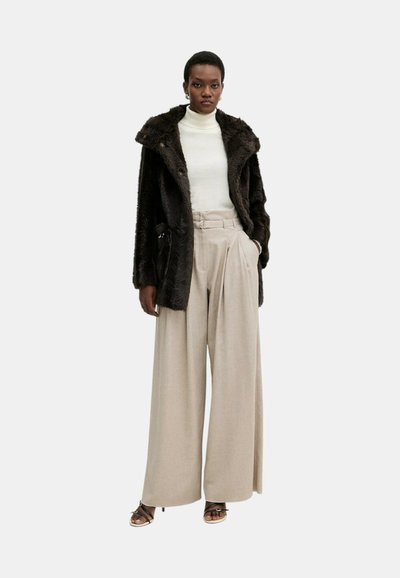 Donna che indossa un cappotto in pelliccia sintetica marrone scuro, dolcevita bianco, pantaloni beige a gamba larga con cintura e tacchi neri con punta aperta, in piedi contro uno sfondo bianco.