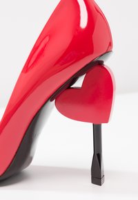 Chaussure à talon haut rouge brillant avec un talon aiguille noir en forme de cœur rouge sur fond blanc.